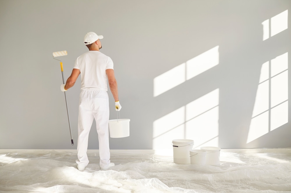 Interior Painting. Back view of a male painter in white clothes holding a bucket and paint roller going painting a grey wall in empty room with copy space. Renovation, apartment repair and renovating house concept.